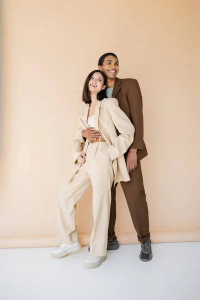 Full length of happy african american man in brown suit embracing stylish asian woman on beige background — Stock Photo