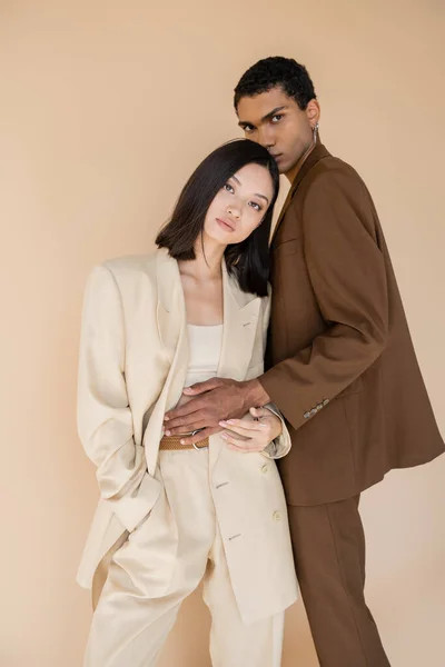 African american guy in brown blazer hugging asian woman in stylish suit isolated on beige — Stock Photo