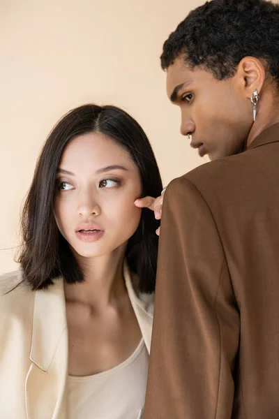 Morena asiático mujer mirando lejos cerca africano americano hombre en marrón chaqueta aislado en beige - foto de stock