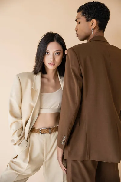 Trendy asian woman in ivory pantsuit standing with hand in pocket near african american man in brown blazer isolated on beige — Stock Photo