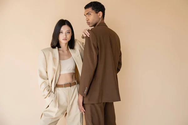 Mujer asiática en traje de pantalón de moda mirando a la cámara mientras posa con la mano en el bolsillo cerca de hombre afroamericano aislado en beige - foto de stock