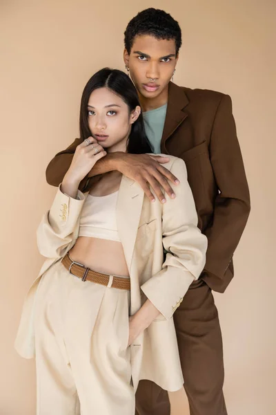 Hombre afroamericano de moda abrazando a mujer asiática en traje de pantalón y mirando a la cámara aislada en beige - foto de stock