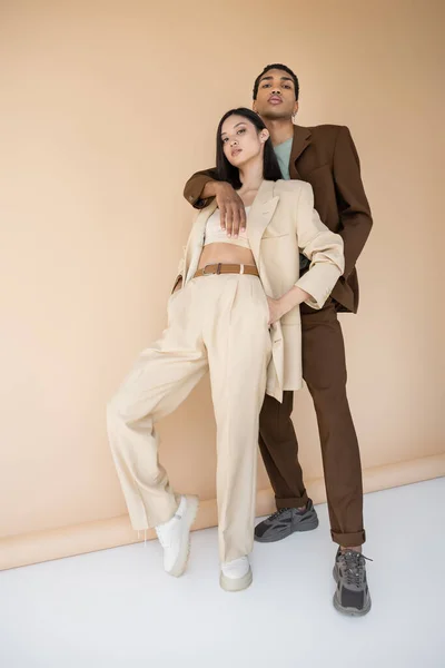 Full length of african american man hugging trendy asian model in ivory pantsuit on beige background — Stock Photo
