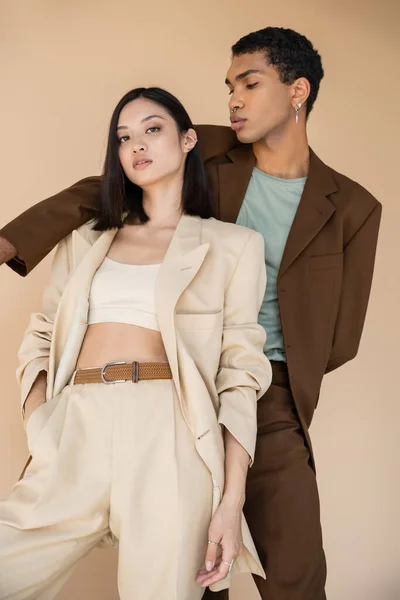 Sensual asian woman in ivory suit posing with hand in pocket near african american man in brown blazer isolated on beige — Stock Photo