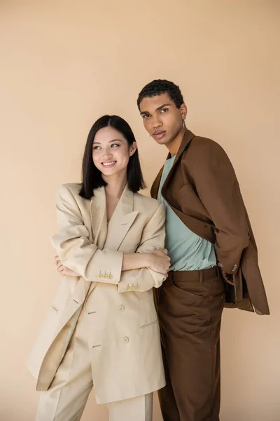 Cheerful asian woman posing with folded arms near african american man in brown suit isolated on beige — Stock Photo