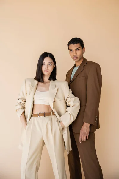 Morena mujer asiática con las manos en los bolsillos mirando a la cámara cerca de moda hombre afroamericano aislado en beige - foto de stock