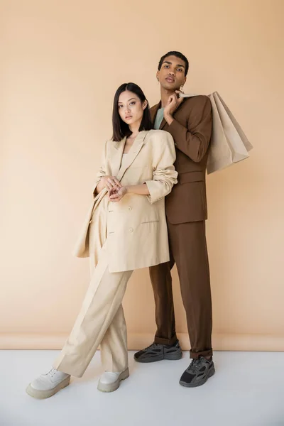 Pleine longueur de femme asiatique élégant en costume regardant la caméra près de l'homme afro-américain avec des sacs à provisions sur fond beige — Photo de stock