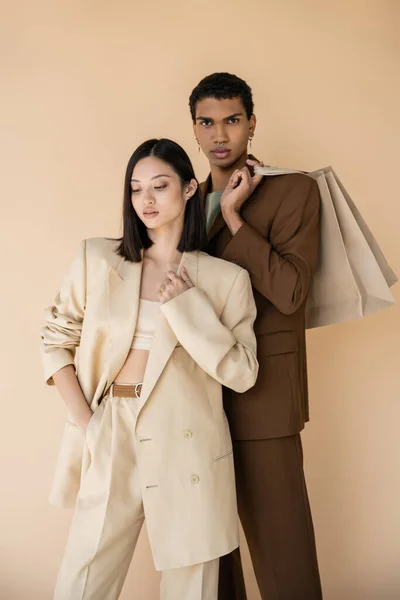 Africano americano homem com compras sacos olhando para câmera perto asiático mulher no pantsuit isolado no bege — Fotografia de Stock