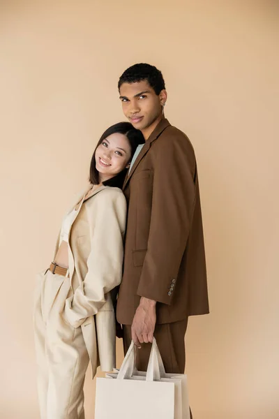 Elegante asiático mujer sonriendo cerca africano americano hombre con compras bolsas aislado en beige - foto de stock
