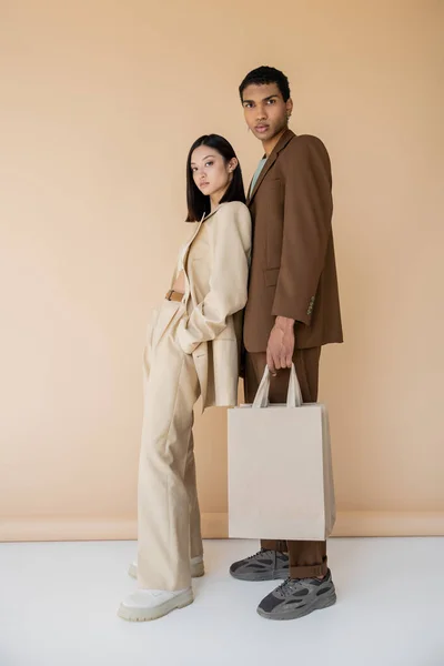 Full length of asian woman in pantsuit posing with hand in pocket near african american guy with shopping bags on beige background — Stock Photo