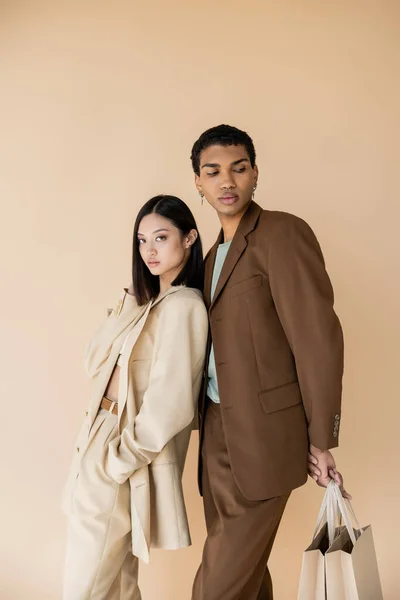 Morena asiático mujer mirando a cámara cerca africano americano hombre con compras bolsas aislado en beige - foto de stock