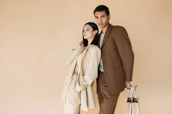 Африканский американец в коричневом костюме позирует с пакетами для покупок рядом с модной азиатской моделью, изолированной на бежевом — стоковое фото