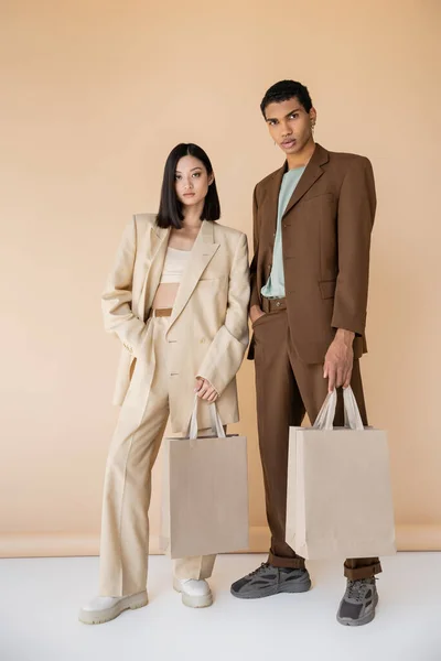 Ganzes interrassisches Paar in trendigen Anzügen posiert mit Einkaufstaschen auf beigem Hintergrund — Stockfoto