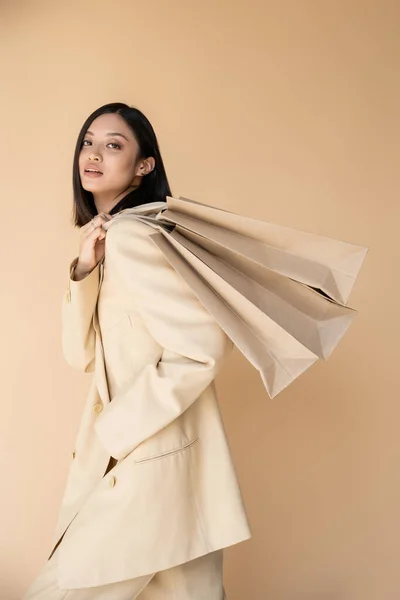 Morena mujer asiática en elegante chaqueta sosteniendo bolsas de compras y mirando a la cámara aislada en beige - foto de stock