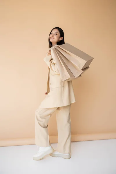 Полная длина веселой азиатской женщины в стильном брючном костюме позирует с пакетами покупок на бежевом фоне — стоковое фото