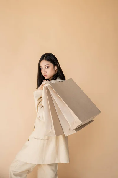 Joven y de moda mujer asiática con bolsas de compras mirando a la cámara aislada en beige - foto de stock