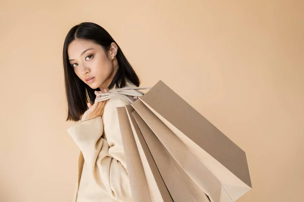 Sensual mujer asiática en chaqueta de marfil sosteniendo bolsas de compras y mirando a la cámara aislada en beige - foto de stock