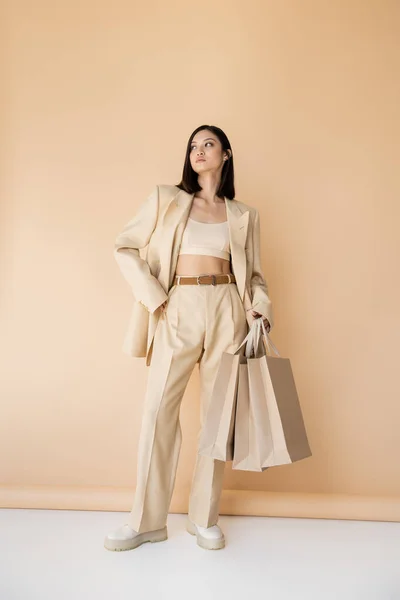 Longitud completa de la mujer asiática con bolsas de compras posando en elegante traje de pantalón de marfil sobre fondo beige - foto de stock