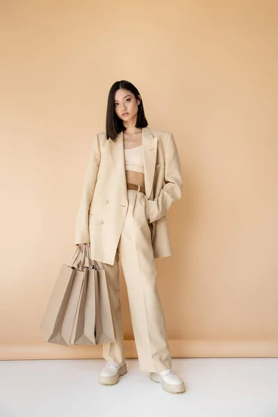 Full length of brunette asian woman in stylish pantsuit standing with shopping bags on beige background — Stock Photo
