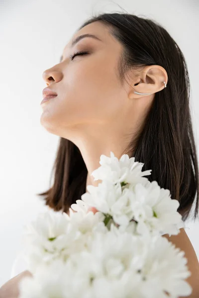 Charming asian woman posing with closed eyes near white fresh flowers isolated on grey — Stock Photo