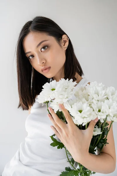 Morena mujer asiática con piel perfecta y maquillaje natural mirando a la cámara cerca de flores blancas aisladas en gris - foto de stock