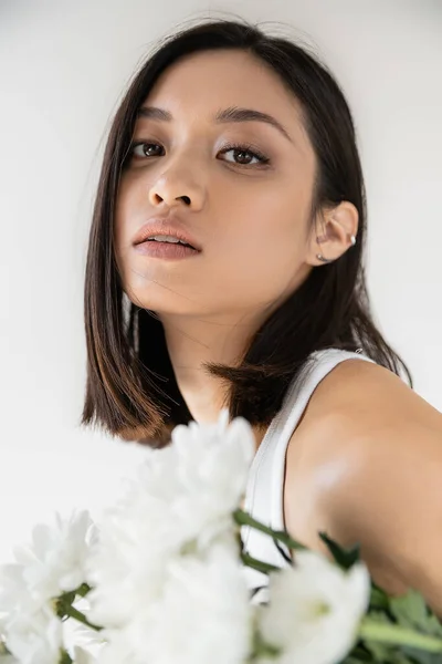 Porträt einer brünetten asiatischen Frau mit sauberer Haut und natürlichem Make-up, die in die Kamera schaut, in der Nähe weißer Blumen isoliert auf grau — Stockfoto