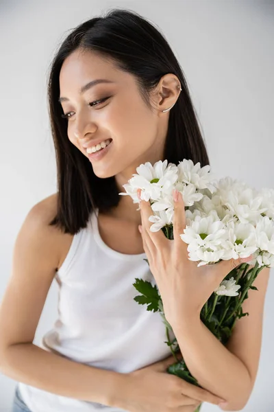 Morena mujer asiática con pendiente de puño sonriendo cerca de flores blancas aisladas en gris - foto de stock