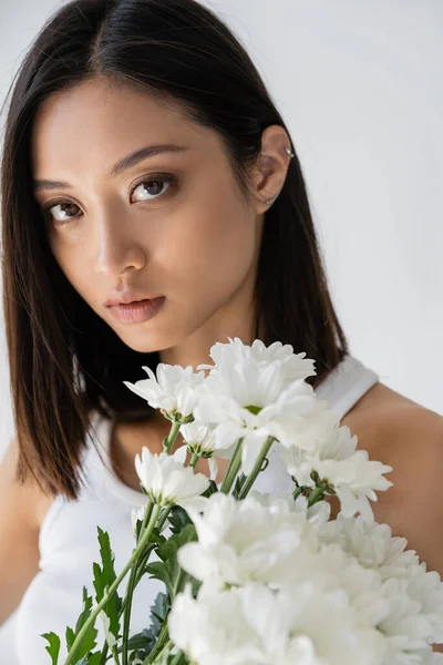 Portrait of sensual asian woman with brunette hair and natural makeup near white flowers isolated on grey — Stock Photo