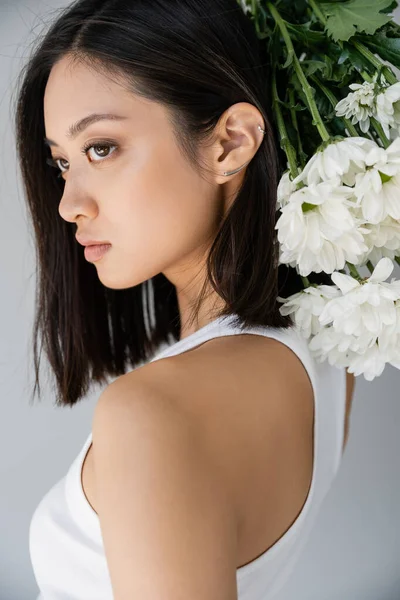 Joven mujer asiática con pelo morena y piel perfecta mirando hacia otro lado cerca de crisantemos blancos aislados en gris - foto de stock