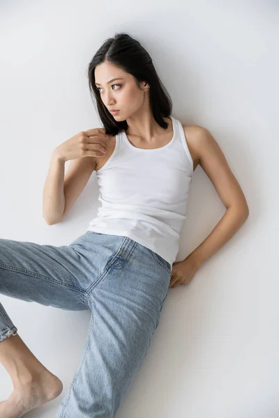 Vue grand angle de femme asiatique en débardeur blanc et jeans regardant loin sur fond gris — Photo de stock