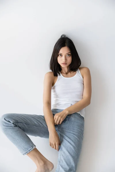 Brünette asiatische Frau in blauen Jeans und weißem Tank-Top sitzt und schaut in die Kamera auf grauem Hintergrund — Stockfoto
