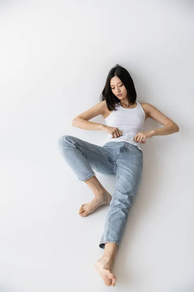 Longitud completa de descalzo asiático mujer en blanco tanque superior y pantalones vaqueros acostado sobre gris fondo - foto de stock