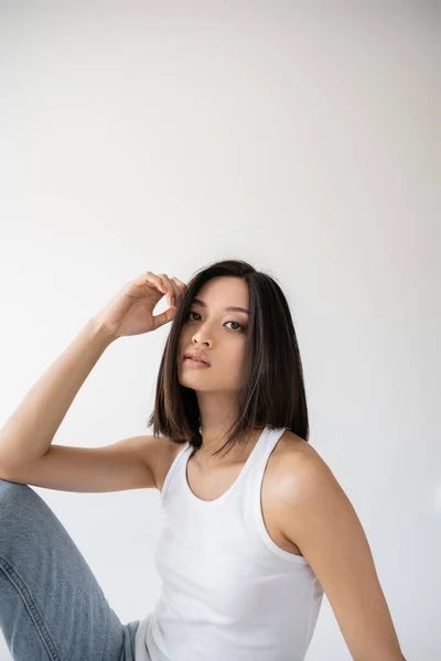 Brunette asian woman in white tank top holding hand near face and looking at camera on grey background — Stock Photo