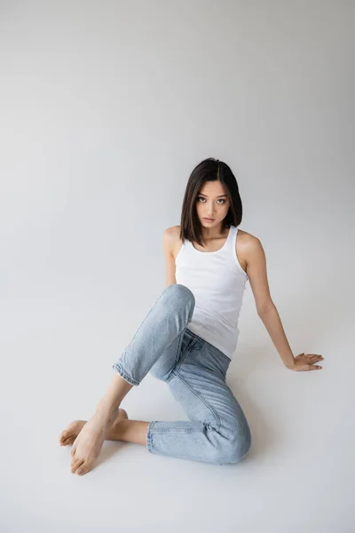 Full length of barefoot asian woman posing in white tank top and blue jeans on grey background — Stock Photo