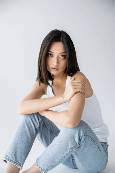Encantador asiático mujer en azul jeans mirando cámara mientras sentado en gris fondo - foto de stock