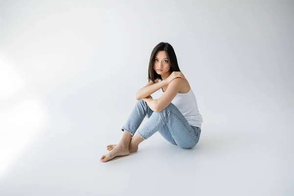 Volle Länge der asiatischen Frau in blauen Jeans und Tank-Top schaut in die Kamera, während sie auf grauem Hintergrund sitzt — Stockfoto