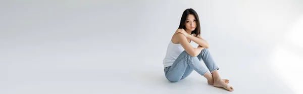 Longitud completa de modelo asiático delgado en pantalones vaqueros azules y camiseta blanca sentado sobre fondo gris, bandera - foto de stock