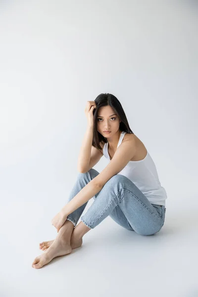 Tutta la lunghezza della donna asiatica a piedi nudi in canotta bianca e jeans seduta e guardando la fotocamera su sfondo grigio — Foto stock