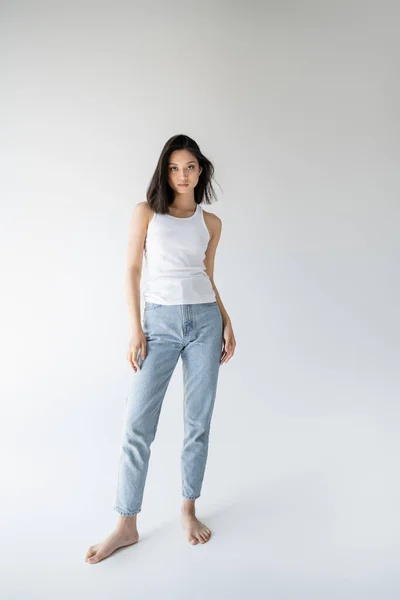 Full length of barefoot and slim asian woman in tank top and jeans standing on grey background — Stock Photo