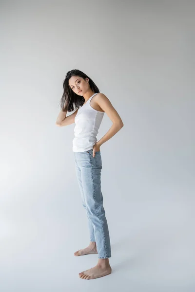 Повна довжина босоніж азіатської моделі в баку зверху позує з рукою в задній кишені блакитних джинсів на сірому фоні — стокове фото