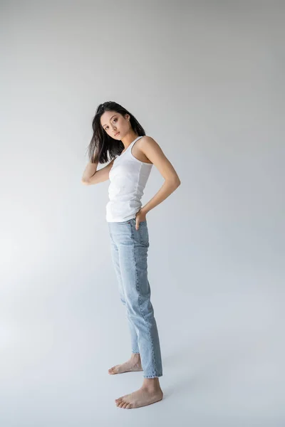 Full length of barefoot asian model in tank top posing with hand in back pocket of blue jeans on grey background — Stock Photo