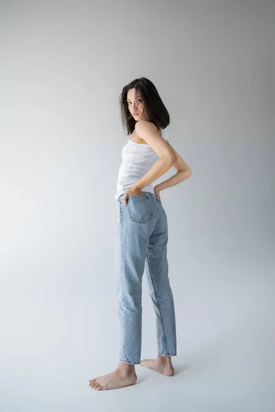 Full length of barefoot asian woman in white tank top and blue jeans holding hands in back pockets on grey background — Stock Photo