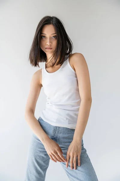Brunette asian woman in white tank top and blue jeans looking at camera isolated on grey — Stock Photo