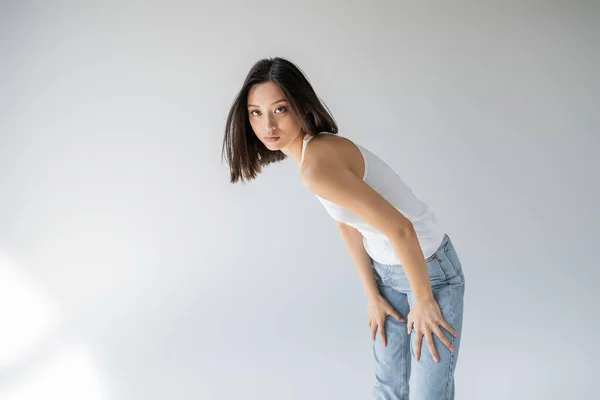 Jovem e delgado asiático mulher no jeans e tanque superior olhando para câmera no cinza fundo — Fotografia de Stock