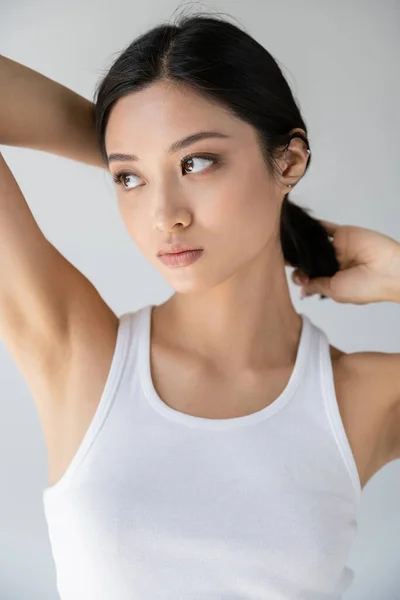 Красива азіатська жінка в білому баку зверху торкається брюнетки волосся і дивиться ізольовано на сірий — стокове фото