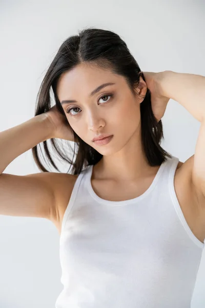 Junge asiatische Frau im weißen Tank-Top berührt brünettes Haar und schaut auf Kamera isoliert auf grau — Stockfoto