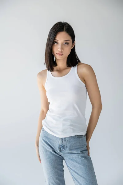 Sinnliche asiatische Frau in Jeans und weißem Tank-Top schaut in die Kamera isoliert auf grau — Stockfoto