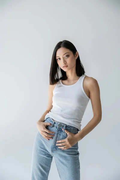 Молода і струнка азіатська модель позує в блакитних джинсах і білий бак зверху ізольована на сірому — стокове фото