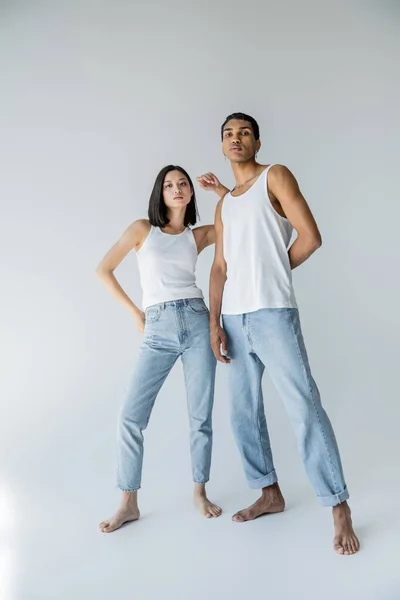 Longitud completa de la pareja interracial descalza en pantalones vaqueros azules y camisetas blancas mirando a la cámara sobre fondo gris - foto de stock