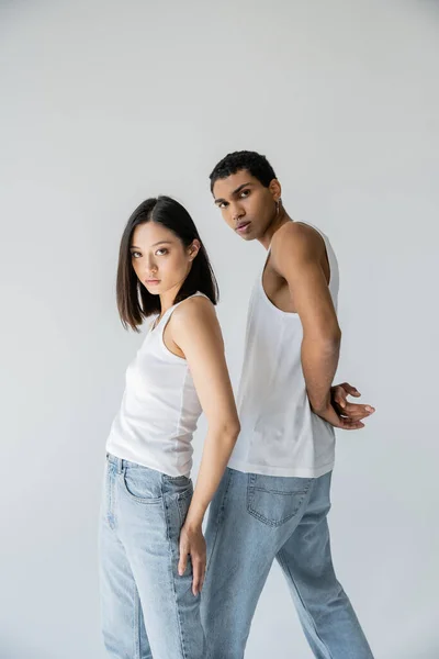 Jovem e moderno casal interracial em tops tanque branco e jeans olhando para a câmera isolada no cinza — Fotografia de Stock